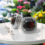 Load image into Gallery viewer, Crimson Allure: Cushion Cut Garnet Stud Earrings with Moissanite Halo.
